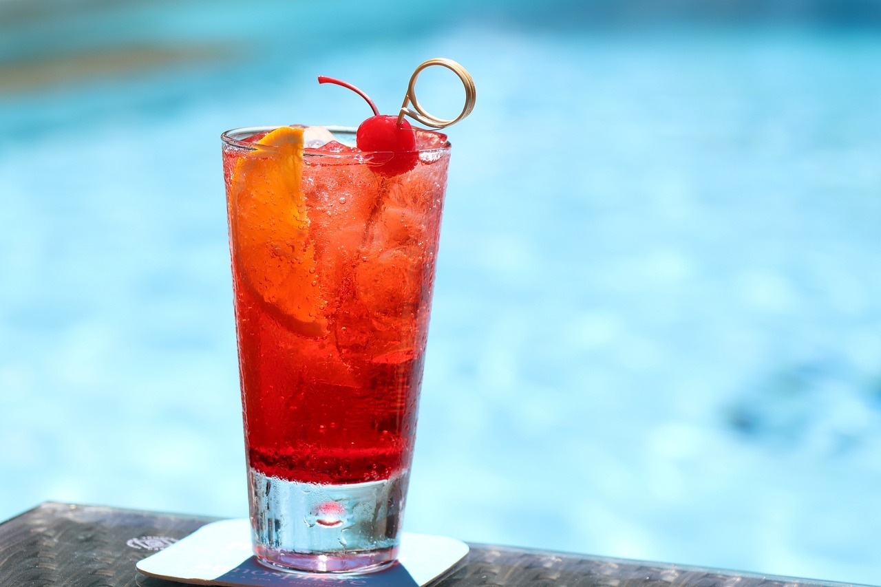 white sand, red cocktail, transparent straw, cocktail glass on the sand, blue sky, clouds, aquamarine water